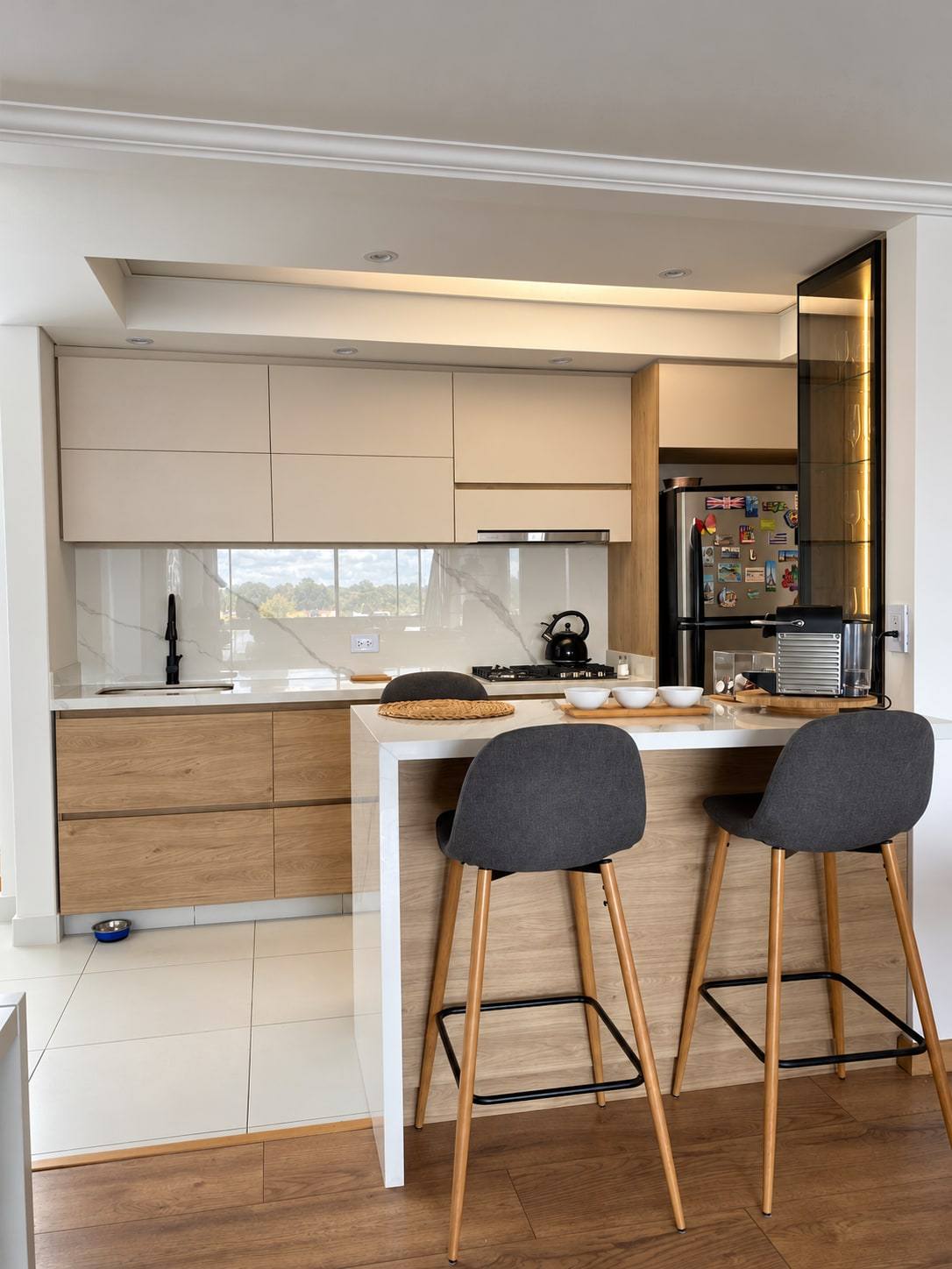 Cocina con muebles bajos en madera, muebles altos claros y barra blanca con frente de madera y dos taburetes.